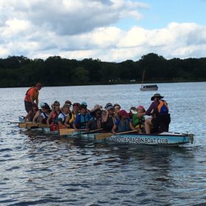 Image of full dragon boat with drummer and steersperson coming in close to shore. 