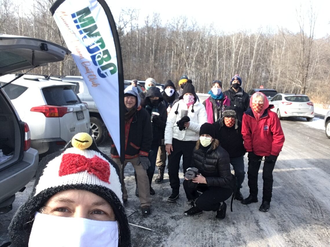 MNDBC Team members pose by our new teardrop banner with our logo as we enjoy beverages in the parking lot of the Richardson Nature Center in Bloomington MN. Packed snow covers the pavement, and leafless trees are in the background.