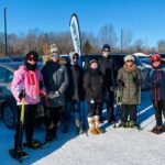 Seven members of the Minnesota Dragon Boat Club standing near a club banner in a parking lot at Hyland Lake Park Reserve in Bloomington. Snow is on the ground and leafless trees and shrubs are in the background. The sky is blue, showing through tree branches.