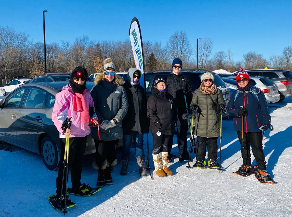 Seven members of the Minnesota Dragon Boat Club standing near a club banner in a parking lot at Hyland Lake Park Reserve in Bloomington. Snow is on the ground and leafless trees and shrubs are in the background. The sky is blue, showing through tree branches.