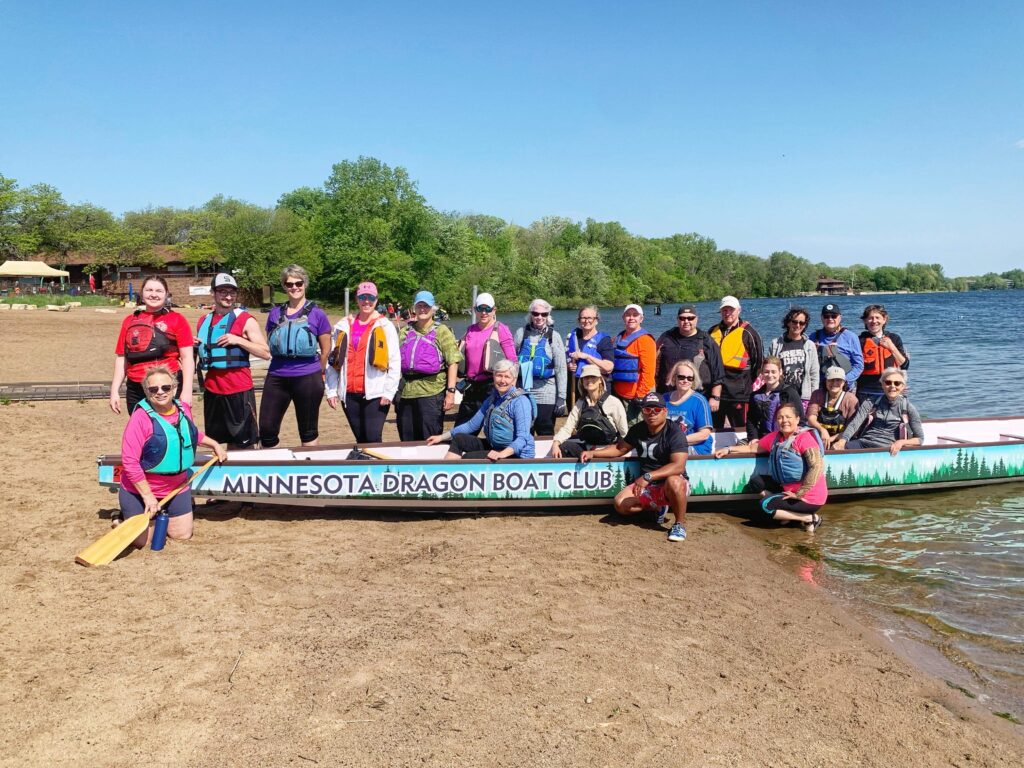 MNDBC team members pose by our boat, Aurora, at Lake Phalen Regional Park's beach. Part of the boat is up on the sand beach, part in the lake. Blue water and trees behind the boat, blue skies.