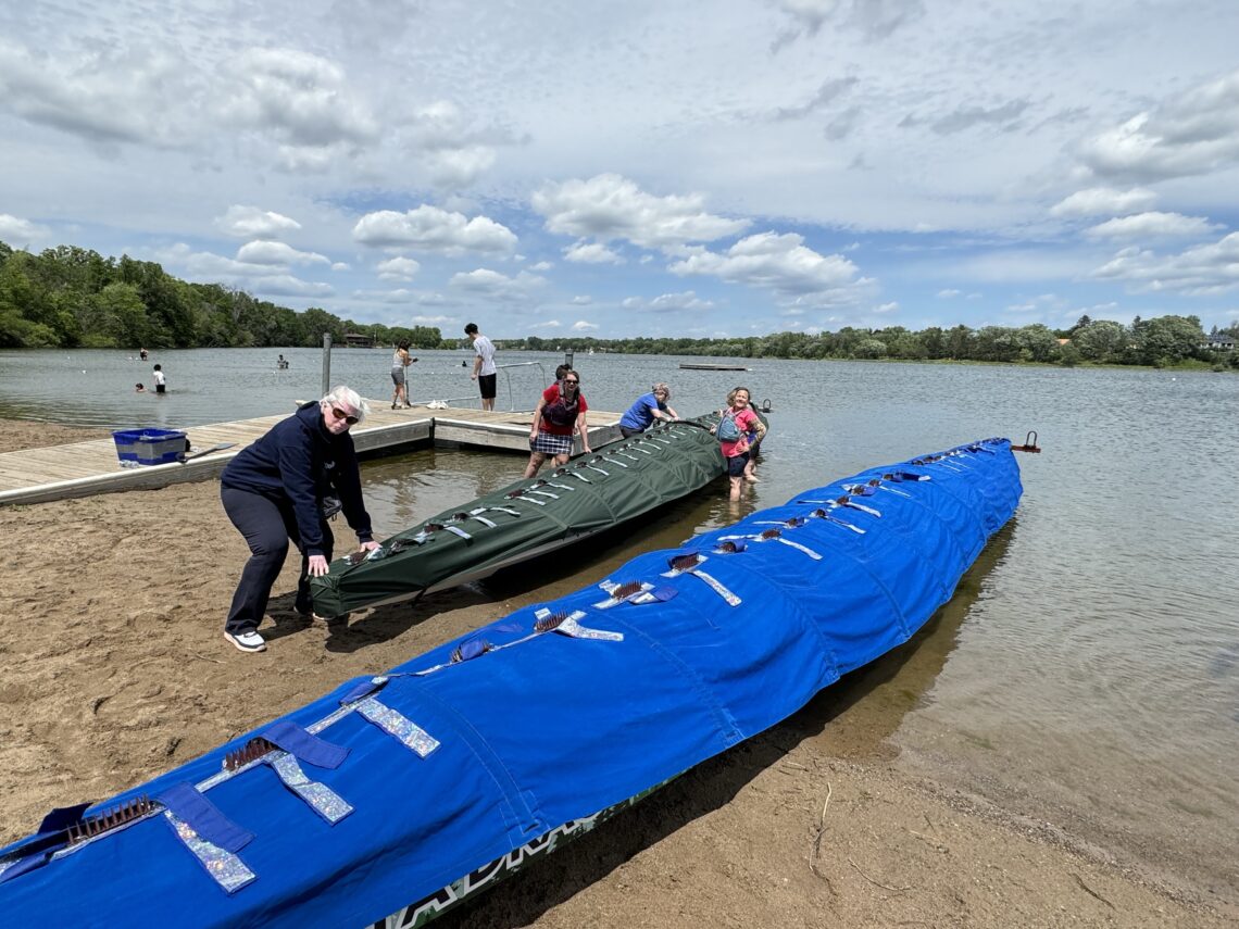 Two dragon boats along a beach, one with a blue cover and one with a green cover. A few people are standing around the boat with the green cover.