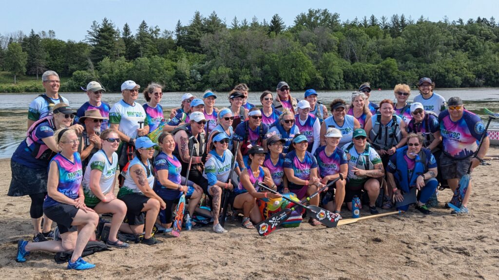 The combined MNDBC Teams Blue and ACP pose for a photo on the sandy beach in front of Badger Lake at Kennedy Memorial Park. The lake is in the background with a backdrop of trees and blue sky along the horizon.
