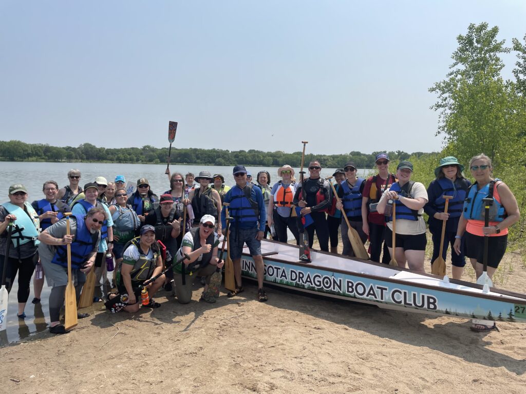 Photo of our Learn to Paddle graduates. People are standing on a standing beach around the bow of a dragon boat. Lake Phalen is in the background, with trees on the horizon and blue skies.
